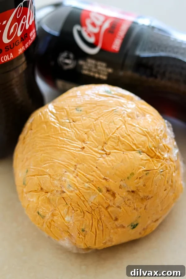 A Buffalo Chicken Cheese Ball tightly wrapped in plastic wrap, resting beside a bottle of Coca-Cola Zero Sugar, ready for chilling.