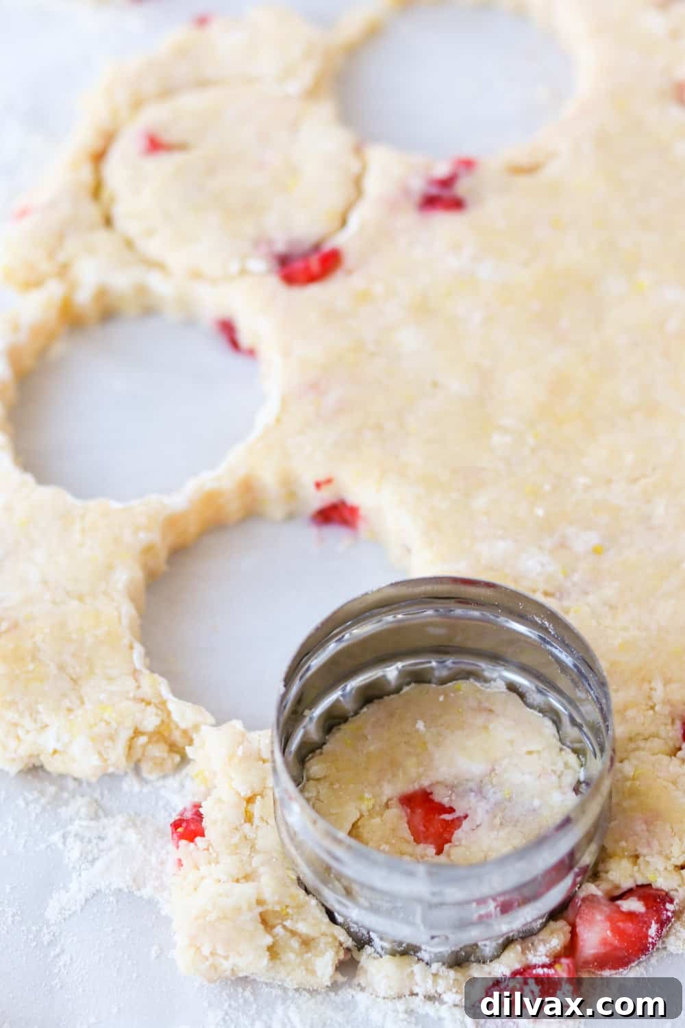 A round cookie cutter in action, pressing through scone dough filled with visible strawberry pieces, ready to be transferred to a baking sheet.