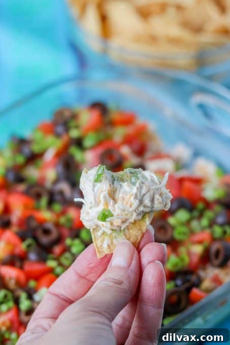 A hand holding a thick tortilla chip, scooping up a generous amount of 7-Layer Dip.