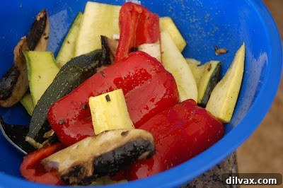 Various sliced raw vegetables, including zucchini, yellow squash, red bell pepper, Portobello mushrooms, and red onion, being gently tossed in a large bowl with the olive oil and seasoning mixture.