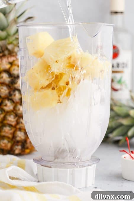 Ice, coconut rum, and fresh pineapple chunks being added to a blender for making a tropical rum slush cocktail.