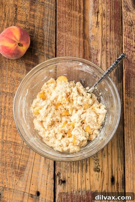 A close-up shot of scone dough with visible chunks of fresh peaches being mixed with a fork.