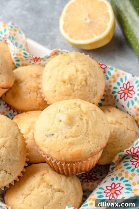 A stack of golden-brown Lemon Zucchini Muffins on a white plate, with a scattering of lemon zest and a whole lemon nearby.