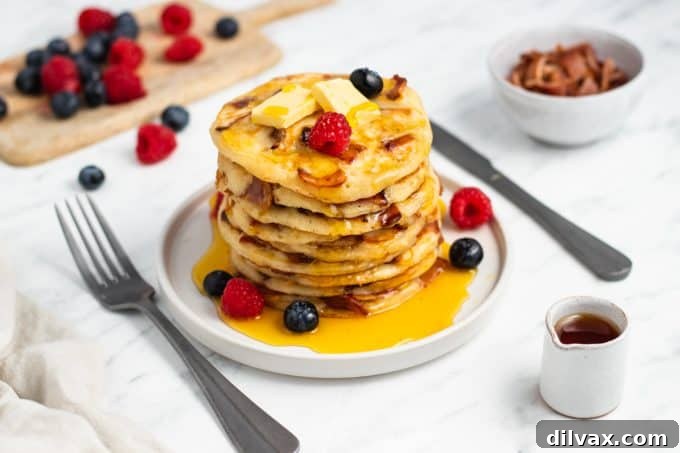 A stack of golden brown buttermilk bacon pancakes, garnished with a pat of butter and a drizzle of maple syrup.