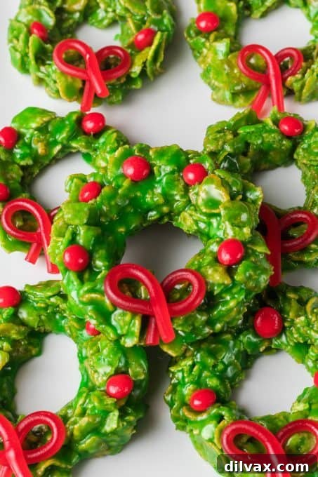 A close-up of green cornflake cookies, featuring a red licorice bow and scattered red hot cinnamon candies.