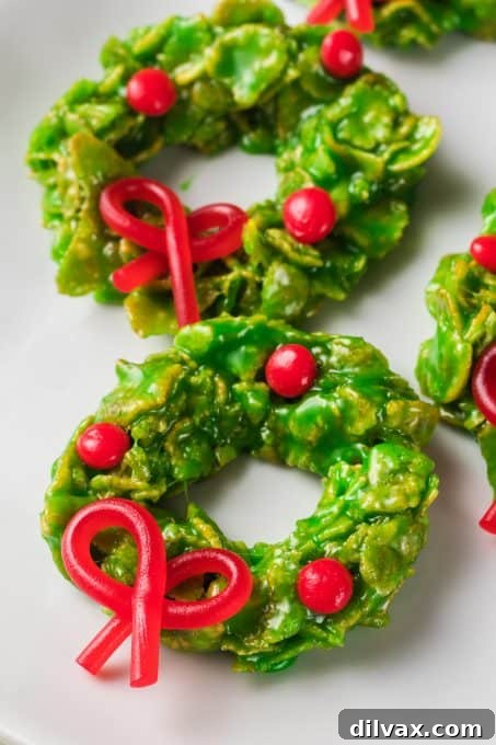 A vibrant display of green Cornflake Wreath Cookies, adorned with red licorice bows and cinnamon candies, presented on a white plate.