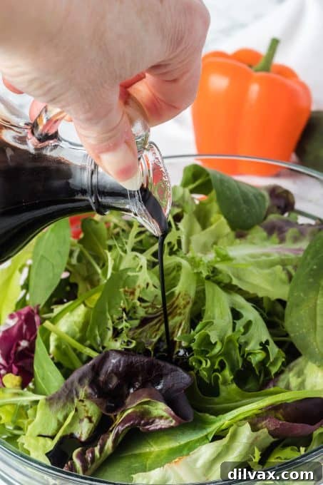 A hand expertly drizzling a rich, dark balsamic reduction from a glass cruet over a vibrant green salad, making it a perfect finish.