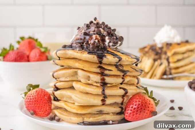 A tall stack of fluffy homemade chocolate chip pancakes, drizzled with syrup and topped with whipped cream.