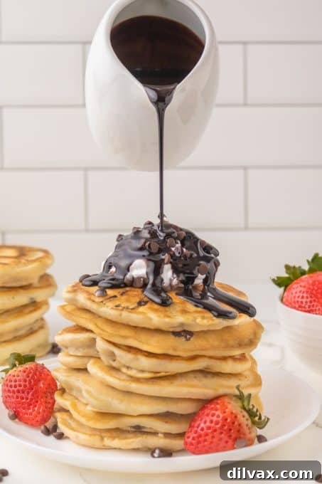 Chocolate sauce being poured over a stack of fluffy chocolate chip pancakes.