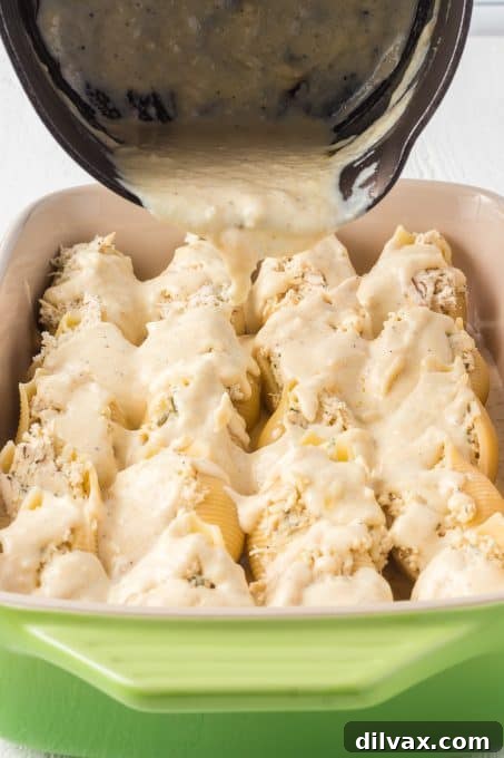 Pouring Garlic Alfredo Sauce over chicken stuffed shells arranged in a baking dish.