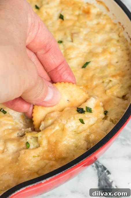 Someone dipping a cracker into a bowl of creamy baked artichoke dip.