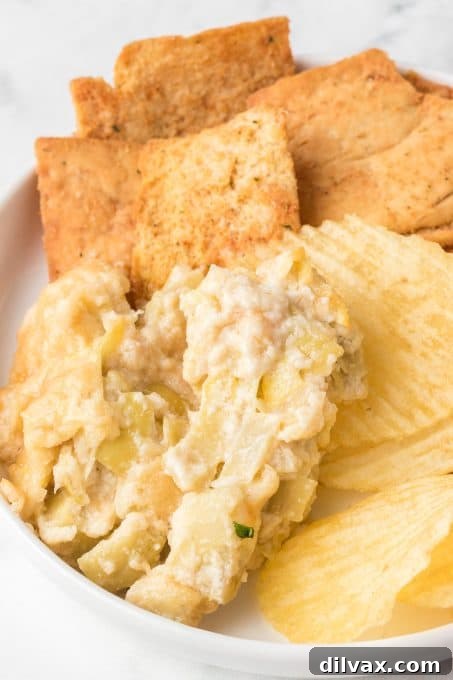 An array of chips, crackers, and vegetable sticks surrounding a bowl of baked artichoke dip.