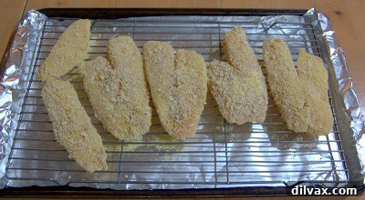 Several breaded tilapia fillets neatly arranged on a wire rack on a baking sheet, ready for the oven.