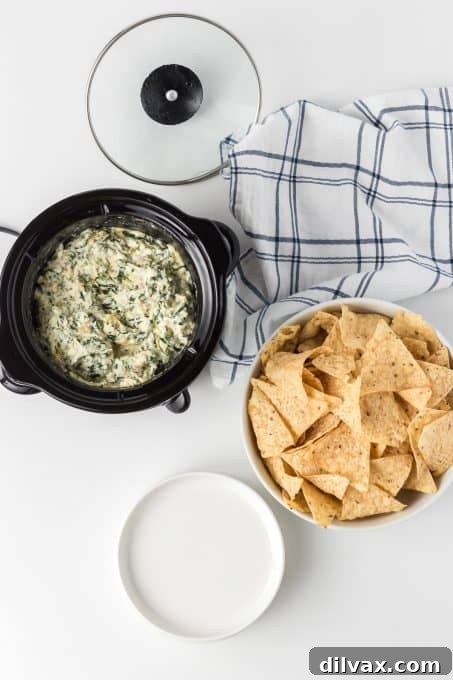 Warm & Cheesy Slow Cooker Spinach Artichoke Dip 6 A slow cooker filled with warm spinach dip and artichokes, surrounded by an array of tortilla chips and fresh vegetables for dipping.