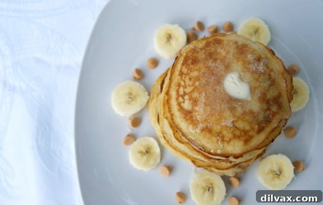 A close-up shot of several golden-brown Banana Peanut Butter Chip Pancakes stacked high, with visible peanut butter chips melting into the surface. The pancakes are garnished with fresh banana slices and a generous pour of maple syrup, making for a truly mouthwatering breakfast.
