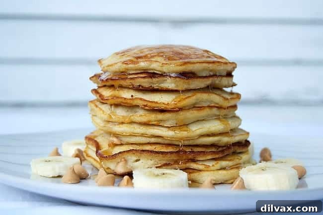 A stack of golden brown Banana Peanut Butter Chip Pancakes, drizzled with maple syrup and topped with fresh banana slices, presented on a rustic wooden board. The melted peanut butter chips are visible, adding to the allure of this comforting breakfast.