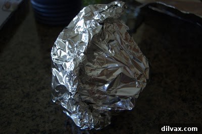 Hands folding the edges of aluminum foil together to completely encase an artichoke.