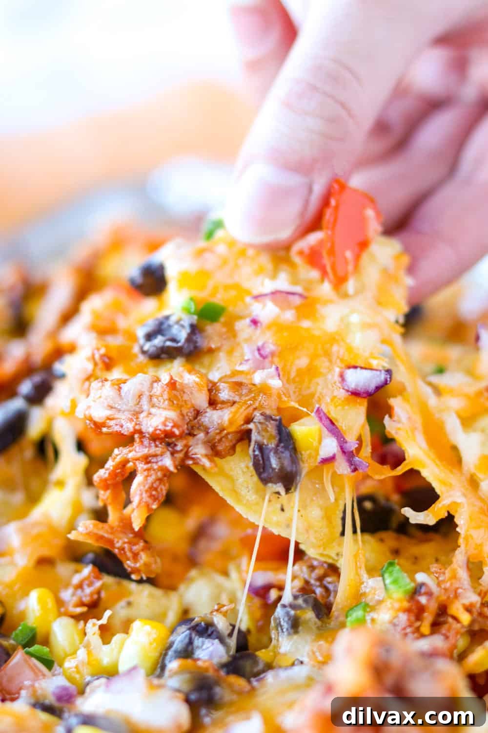 A hand lifting a loaded chicken nacho off a baking tray, full of cheese and toppings.