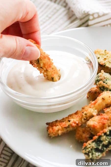 A single oven-baked zucchini stick being dipped into a bowl of creamy Ranch dressing