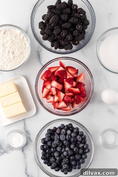Fresh ingredients laid out for making Berry Cobbler: strawberries, blueberries, blackberries, butter, sugar, milk, eggs, flour, baking powder, and salt, ready for preparation.