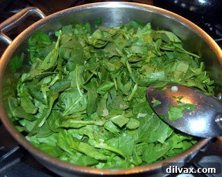 Fresh spinach wilting in a skillet with sautéed onions and garlic, releasing its moisture.
