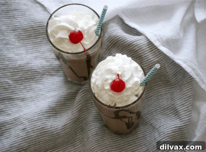 A tall glass filled with a rich Double Chocolate Malted Milkshake, generously topped with whipped cream and a bright red maraschino cherry, ready to be enjoyed on a hot summer day.