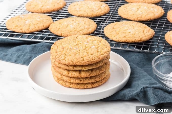 A stack of golden Cornmeal Cookies on a plate, with more cookies cooling on a baking rack in the background.