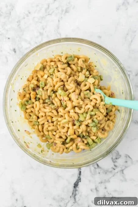A light green spatula mixing cooked macaroni, chopped vegetables, and hard-boiled eggs in a large glass bowl.