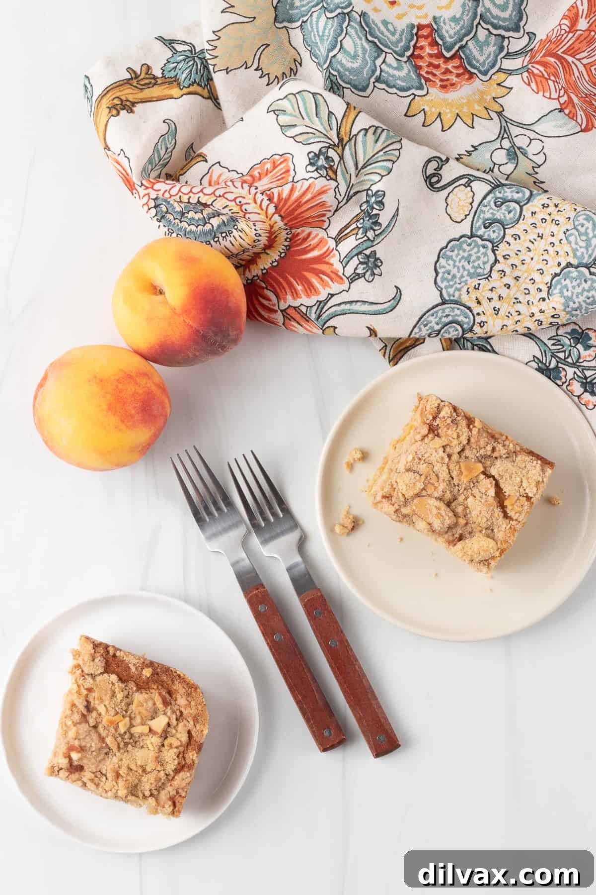 Looking down at two slices of cake on separate plates with two forks, two peaches and a floral napkin.